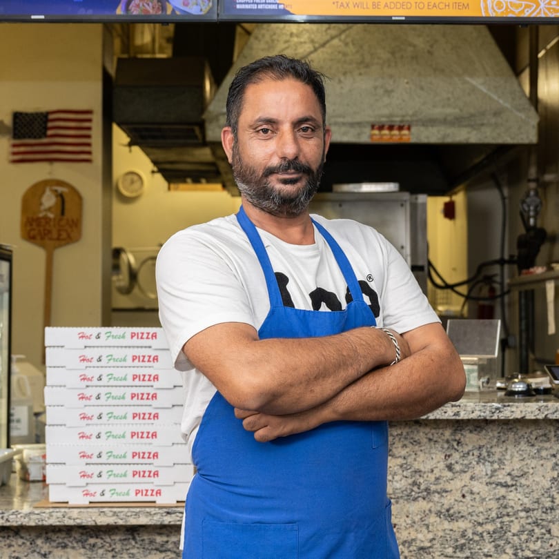Garlex Pizza & Ribs owner in a blue apron, arms crossed, in front of pizza boxes and kitchen.