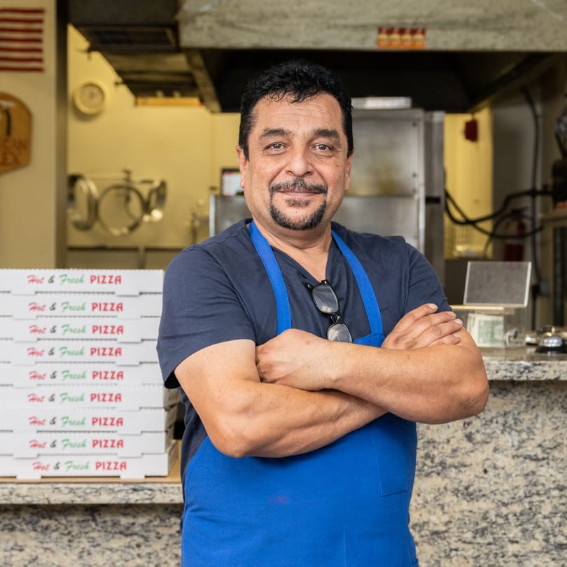 Garlex Pizza & Ribs owner, a man in a blue apron, stands proudly with pizza boxes.