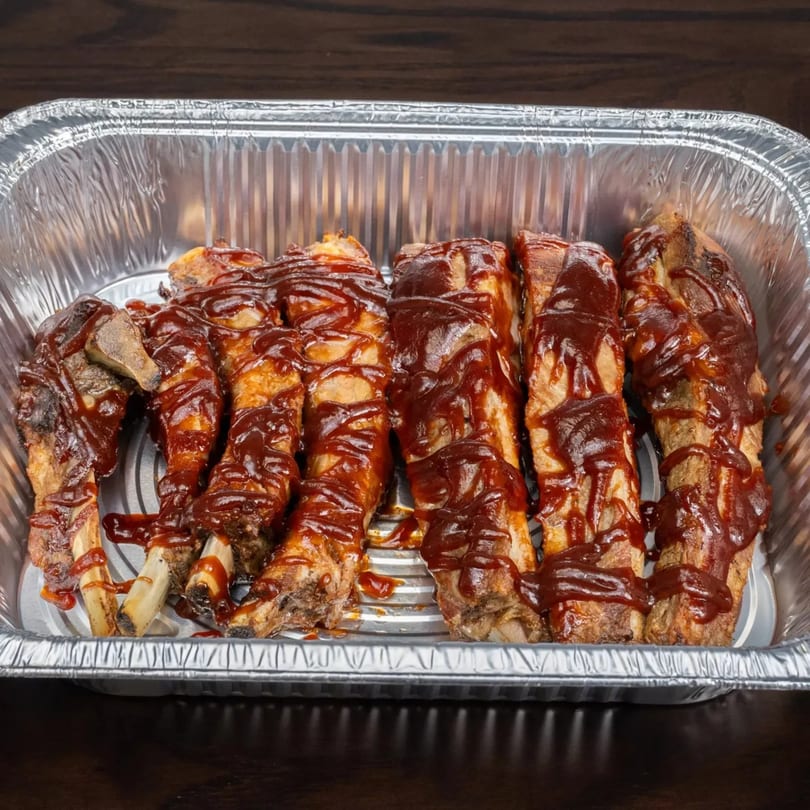 Garlex Pizza & Ribs' BBQ ribs, generously coated in sauce, in a silver foil takeout container.